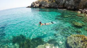 Persona fa snorkeling in una cala rocciosa con acqua cristallina turchese e pesci visibili sotto la superficie